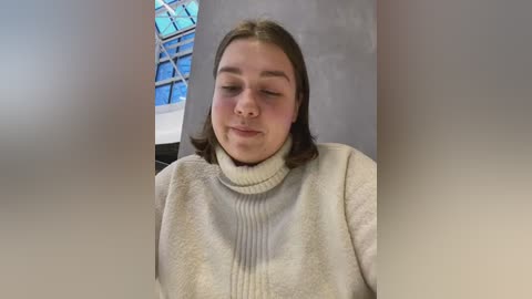 Media: Video of a young woman with light skin and shoulder-length brown hair, wearing a cream-colored turtleneck sweater, sitting in an indoor space with a blue-tiled ceiling and metal structures.