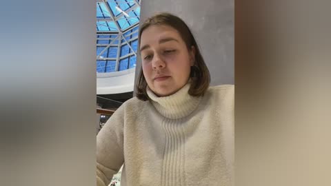 Media: Video of a young woman with light skin and shoulder-length brown hair, wearing a cream-colored turtleneck sweater, standing indoors next to a large glass dome structure.