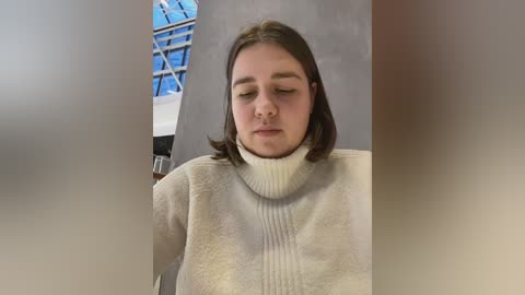 Media: Video of a young woman with shoulder-length brown hair, wearing a cream-colored turtleneck sweater, sitting indoors with a blue-tiled wall and a glass window in the background.