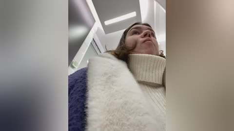 Media: A video of a young woman with light brown hair, wearing a cream-colored turtleneck sweater, standing in an elevator with a fluffy white and blue blanket draped over her shoulders.