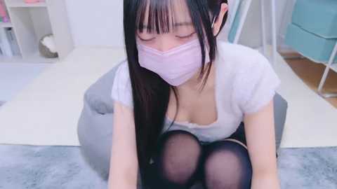 Media: Video of an Asian woman with long black hair, wearing a white top, black tights, and a pink face mask, sitting on a gray beanbag chair in a minimalist room with white walls and light wooden flooring.