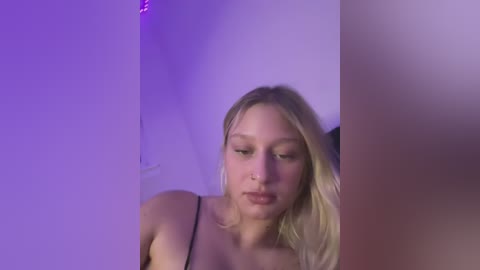 Media: Video of a young blonde woman with fair skin, wearing a black spaghetti strap top, gazing downward. Background features purple and white lighting.