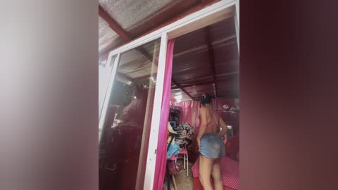 Media: Video of a naked woman with fair skin and long black hair, exiting a small, pink-tiled room with a reflective mirror, revealing a messy interior with clothes and bags.