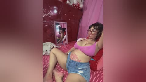 Media: Video of a middle-aged woman with glasses, wearing a pink bra and denim shorts, lying on a red bed with a large mirror reflecting her.