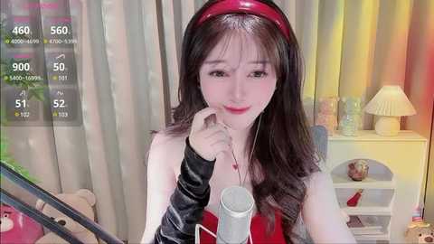 Media: Video of an Asian woman with long black hair, wearing a red headband, a black arm sleeve, and a red dress, holding a microphone, in a brightly lit room with a yellow lamp and a teddy bear.