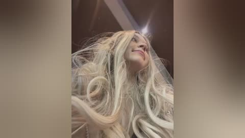 A close-up video of a blonde woman with long, flowing hair, smiling with her eyes closed, set against a blurred, brown background.