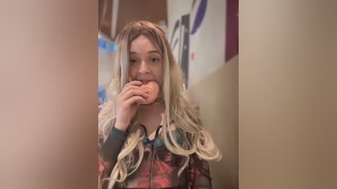 Video of a young woman with long, wavy blonde hair, wearing a floral jacket, eating a sandwich in a dimly lit hallway. Background shows blurred posters and beige walls.