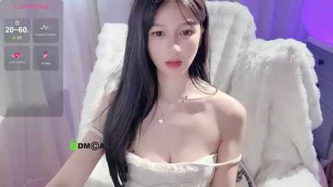 Media: A video of an East Asian woman with long black hair and fair skin, wearing a white strapless dress, sitting on a plush white chair.