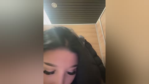 Media: Video of a young Asian woman with long black hair, wearing black clothing, leaning forward in a dimly lit, beige-tiled bathroom with a black ceiling. Her face is partially obscured by the angle.