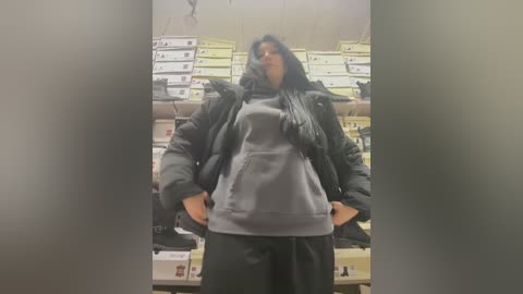 Media: Video of a person wearing a black puffer jacket and gray hoodie, with a scarf, in a store with stacks of boxes in the background.