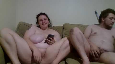 Media: Video of two nude, overweight people with pale skin, sitting on a beige couch, one holding a phone, the other resting with legs spread.