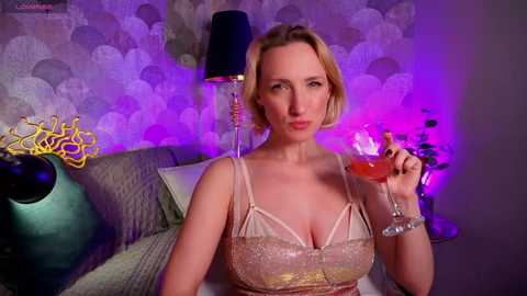 Video of a blonde woman in a pink sequined dress, holding a glass of wine, against a purple and silver-patterned wallpaper background.