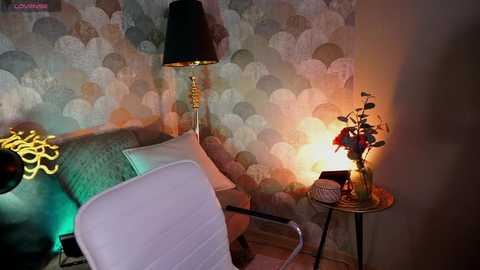 Media: Video of a cozy, vintage-style room with a floral-patterned wallpaper, a beige armchair, a round side table, and a lit lamp emitting a warm glow.