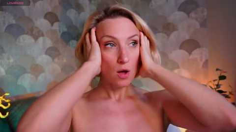 Video of a topless, fair-skinned woman with blonde hair, touching her face, in a room with patterned wallpaper and a green leafy plant in the background.