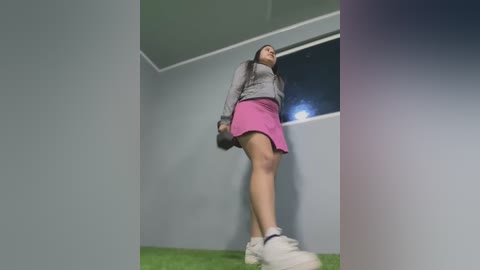 Media: Video of a woman in a sports setting, wearing a grey top, pink skirt, white sneakers, and holding a golf club, with a green carpeted floor and a window in the background.