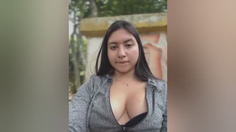 Media: Video of a Latina woman with long black hair, wearing a gray hoodie that exposes her large breasts, standing in front of a rustic, weathered building.