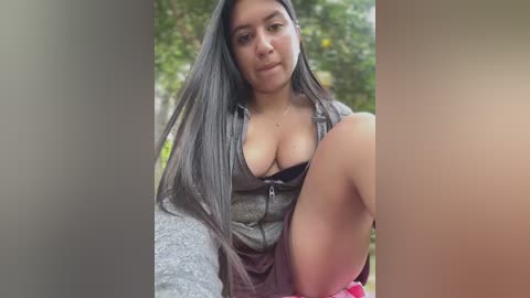 Media: Video of a young woman with long, dark hair, wearing a black top that reveals cleavage, sitting outdoors with blurred greenery background.