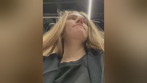 Video of a Caucasian woman with long, blonde hair, wearing a black jacket, reclined in a car with the windows down, hair blowing in the wind.