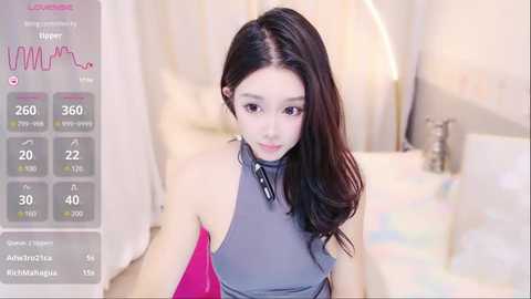 Media: A video of a young Asian woman with long dark hair, wearing a sleeveless grey top, seated in a brightly lit bedroom. A digital health monitor overlay displays her vital signs.