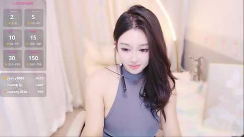 Media: Video of an Asian woman with long, dark hair, fair skin, and a slender physique. She wears a gray, high-necked dress, sitting in a brightly lit, modern bedroom with soft furnishings.