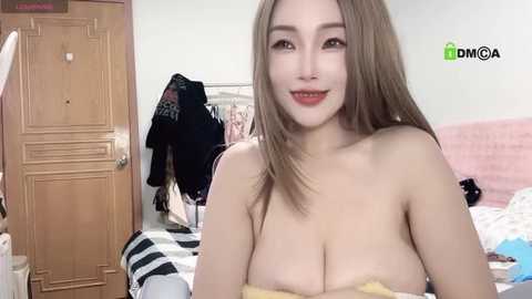 Media: A video of a young East Asian woman with long, straight brown hair, fair skin, and large breasts, smiling in a bedroom with wooden furniture and a bed.