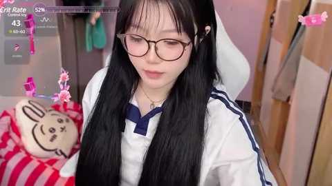 Media: Video of a young East Asian woman with long black hair and glasses, dressed in a white sailor outfit with a blue necktie, sitting at a desk with a plush toy.