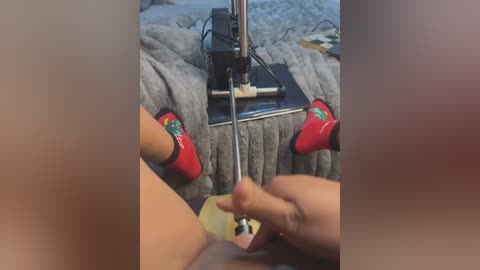 Media: A video of a sewing machine with a light-skinned hand in red socks sewing fabric, on a grey blanket, in a cozy room with a patterned rug.