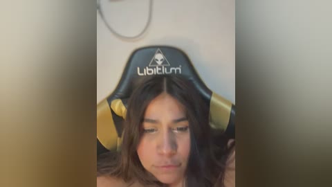 Media: Video of a woman with long, wavy dark hair, wearing a black and gold \"Lubitum\" gaming chair, with a blurred background.