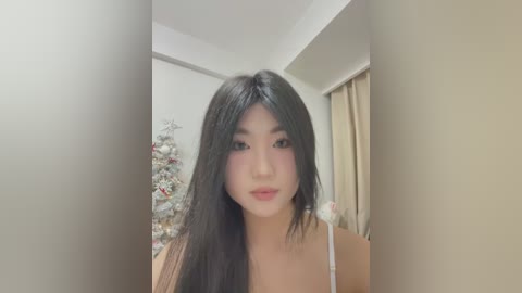 Media: Video of an East Asian woman with long, straight black hair, fair skin, and light makeup, wearing a white top. She stands in a softly lit room with a decorated Christmas tree in the background.