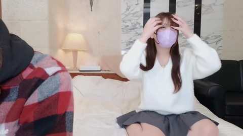 Media: Video of an Asian woman in a white sweater, gray skirt, and lavender mask, sitting on a bed, holding her head, with a red and black plaid blanket draped on a chair. Background features a lamp and a decorative wall.