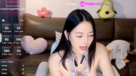 Media: Video of an East Asian woman with long black hair, wearing a revealing white lace top, sitting on a brown leather couch. Background includes plush toys and a pink star pillow.