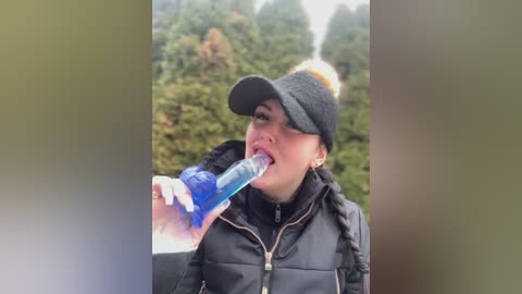 Media: Video of a woman with light skin, wearing a black cap and jacket, drinking from a blue water bottle outdoors in a wooded area.