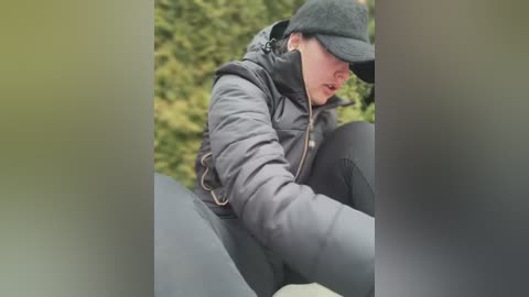 Media: Video of a man in a black flat cap, puffer jacket, and dark pants, crouching, looking down, with blurred greenery in the background.