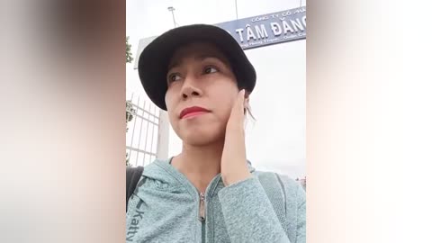 Media: Video of a young woman with medium brown skin, wearing a black hat and light gray hoodie, leaning on a white railing, with a \"TAM BAN\" sign in the background.