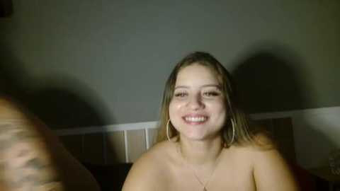 Media: Video of a smiling, topless, light-skinned woman with medium-length blonde hair, wearing a gold necklace, in a dimly-lit room with beige walls.