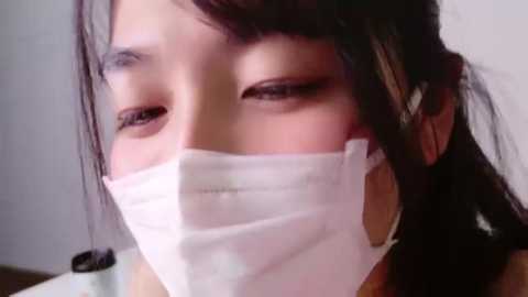 Media: Video of a young East Asian woman with fair skin, wearing a white surgical mask, looking down with eyes half-closed. She has dark hair pulled back and is wearing a black top.