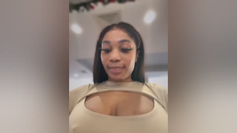 Media: Video of a smiling Black woman with long dark hair, wearing a tight beige top that reveals cleavage. Background features blurred holiday decorations.