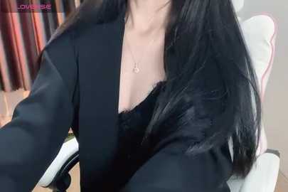 Media: A close-up video of an Asian woman with long, straight black hair, wearing a black blazer over a low-cut black top, revealing cleavage. She sits in a modern white gaming chair.