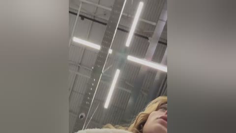 Media: Video of a woman with long blonde hair, partially visible, in a modern office with a high ceiling, fluorescent lights, and white walls.