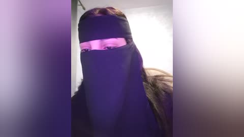 Media: A video of a person wearing a black niqab with a pink tinted section covering the eyes, standing in a dimly lit room with white walls. The image is slightly blurry.