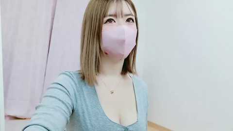 Media: Video of a young Asian woman with shoulder-length light brown hair, wearing a light blue V-neck top, light pink mask, and a delicate necklace. Background shows a light-colored wall and pink curtain.