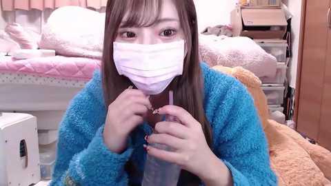 Media: Video of a young East Asian woman in a blue fleece robe, wearing a face mask, holding a syringe in a cluttered bedroom with a bed and storage drawers.