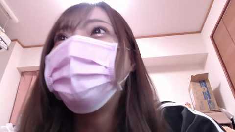 Media: Video of an Asian woman with long, straight brown hair, wearing a white surgical mask, standing in a bright, modern room with light-colored walls, wooden furniture, and an open cardboard box in the background.