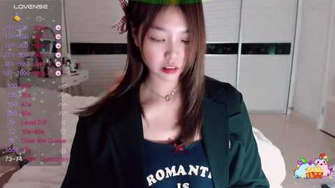 Media: Video of an Asian woman with long brown hair, wearing a green headband, black blazer, and \"ROMANTIC\" T-shirt, indoors with blinds and a table, video chat interface on screen.