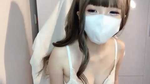 Media: A video of a young Asian woman with long brown hair and bangs, wearing a white face mask, revealing her delicate features. She's in a white tank top, standing in front of a white door.