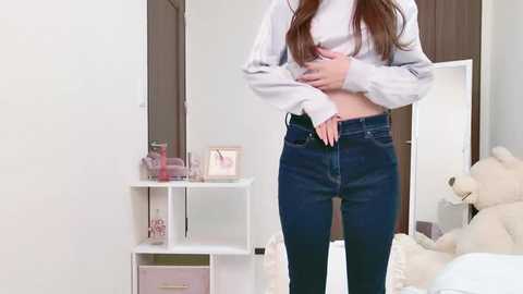 Media: Video of a slim woman with long brown hair, wearing a white blouse and tight dark blue jeans, standing in a modern, minimalist bedroom with a white bed, beige teddy bear, and white shelves.