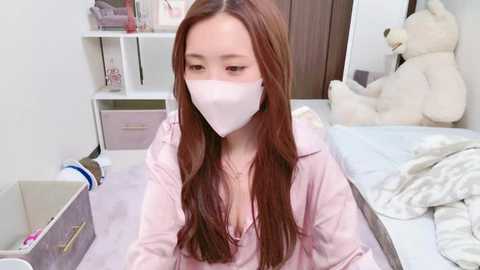 Media: Video of a young Asian woman with long, straight brown hair, wearing a white face mask, pink hoodie, and sitting on a bed in a tidy, white room with a stuffed teddy bear.