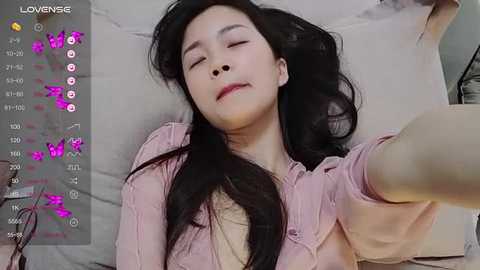 Media: A video of an Asian woman with long black hair, closed eyes, and a pink shirt, lying on a bed, taken from a selfie perspective.