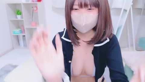 Media: Video of an East Asian woman with a mask, short brown hair, and a black jacket exposing her cleavage. She waves from a white room with shelves and plants.