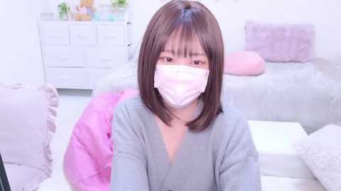 Media: Video of an East Asian woman with shoulder-length dark hair and light skin, wearing a gray robe and pink mask, sitting on a white carpet in a minimalist, pastel-colored room.